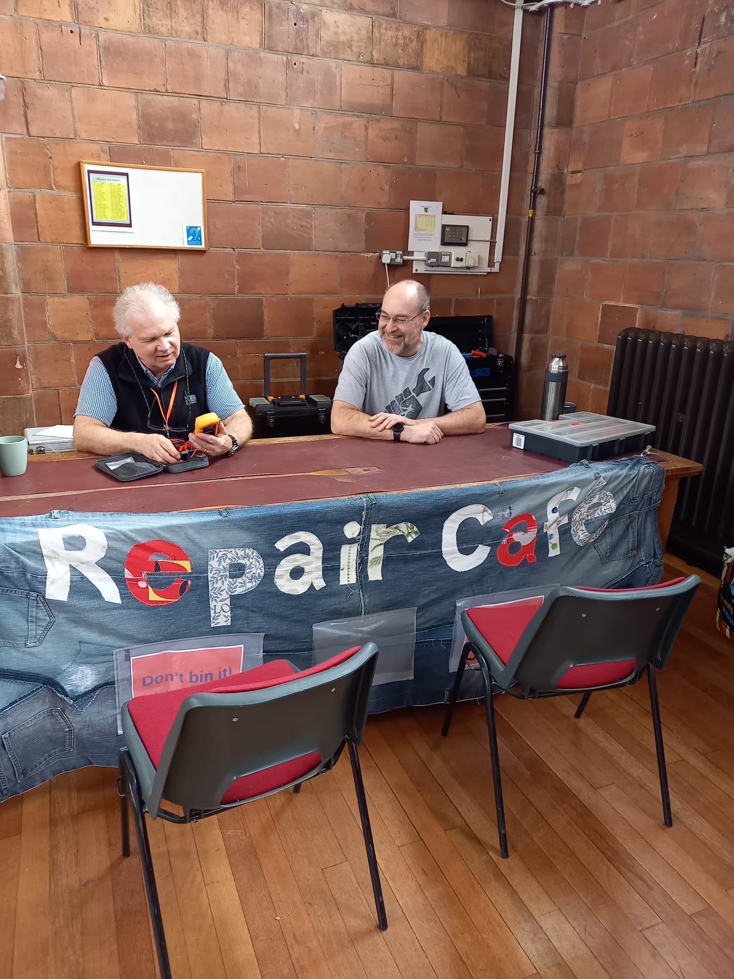 Two men are sitting at a long table with the banner 'repair cafe'. One is inspecting an electrical device
