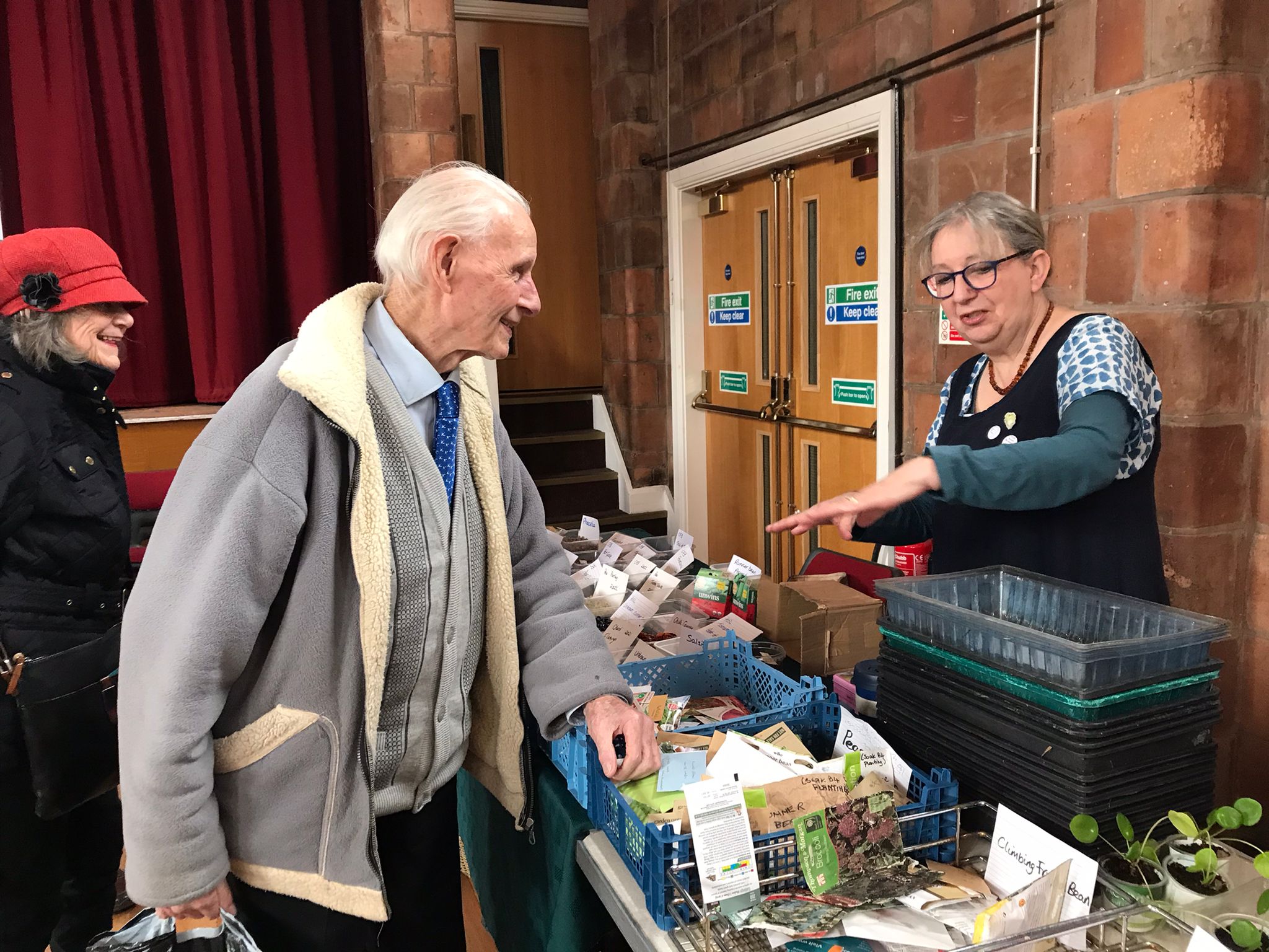 Two older people are talking to a woman who is displaying seeds and seedlings