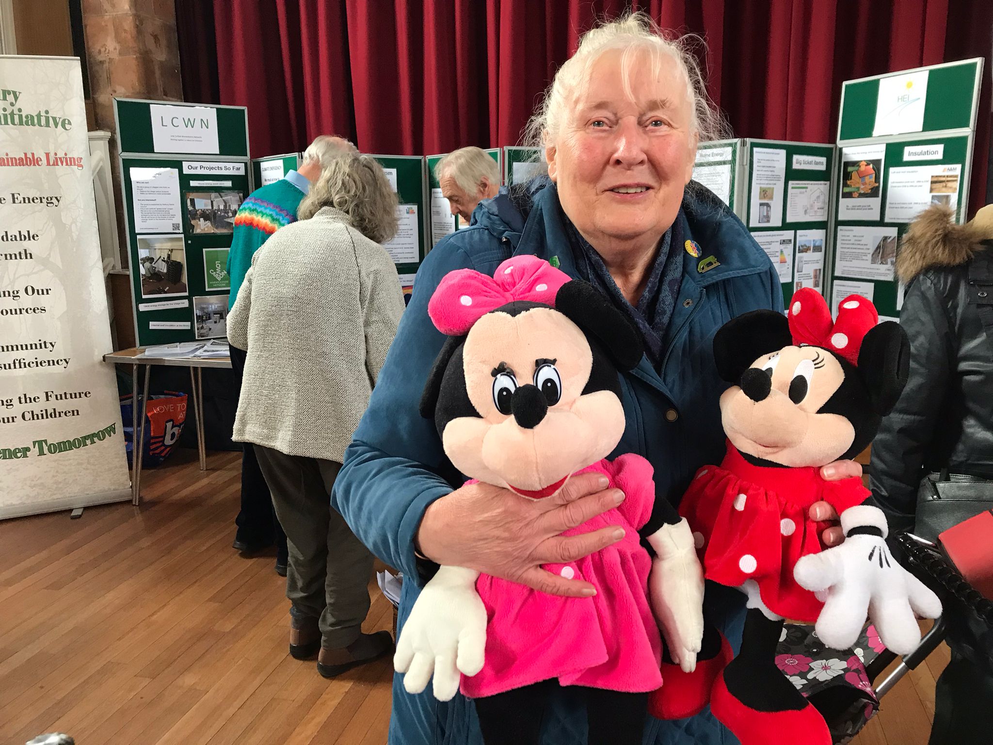 A woman is holding two minnie mouse stuffed toys. She is smiling. behind her is a display for the Low Carbon Warwickshire Network