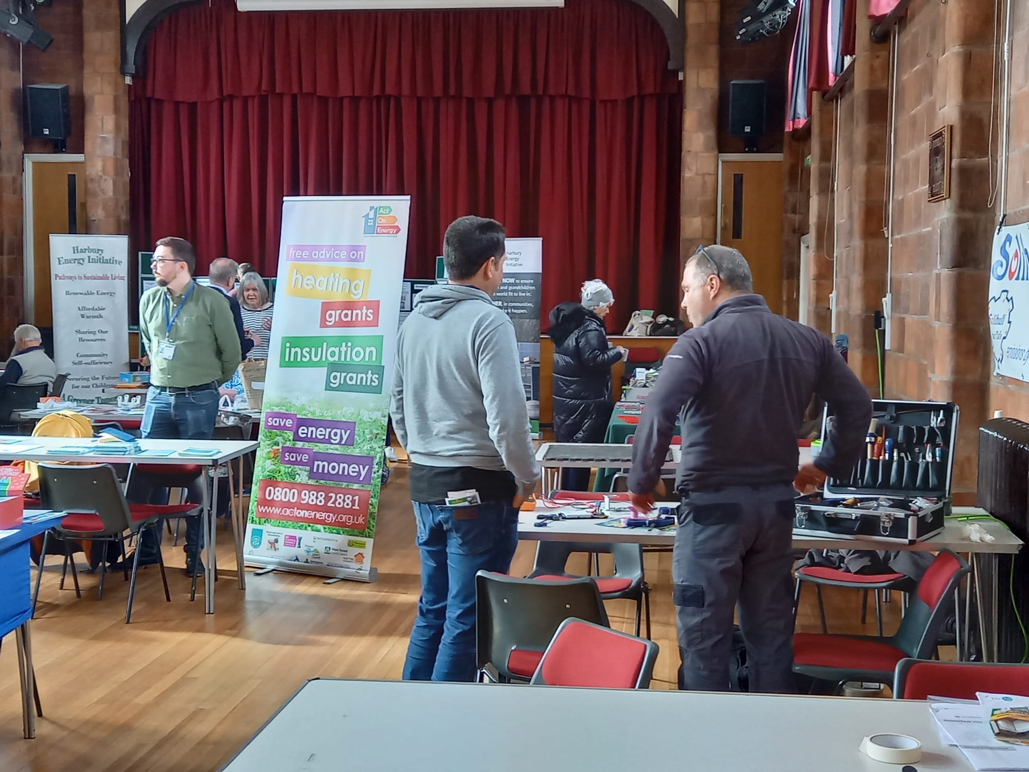 People in a hall with tables with tools and pamphlets. There are posters about saving money and energy. 